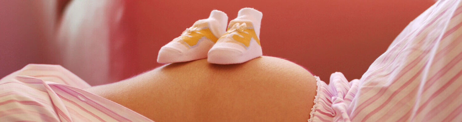 A pair of baby shoes rests on a person's belly, surrounded by soft pink and white fabric.