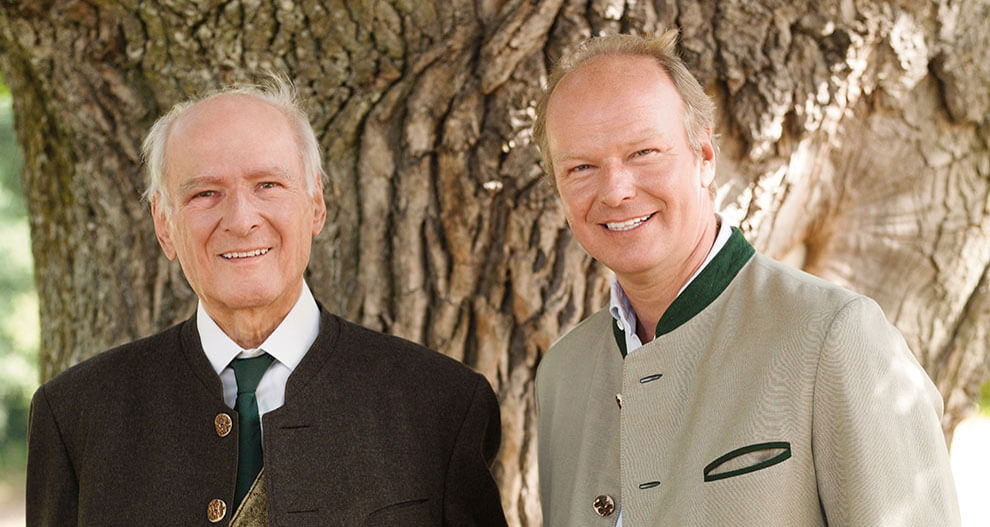 Two men, one older and one younger, dressed in formal attire, stand in front of a tree trunk.