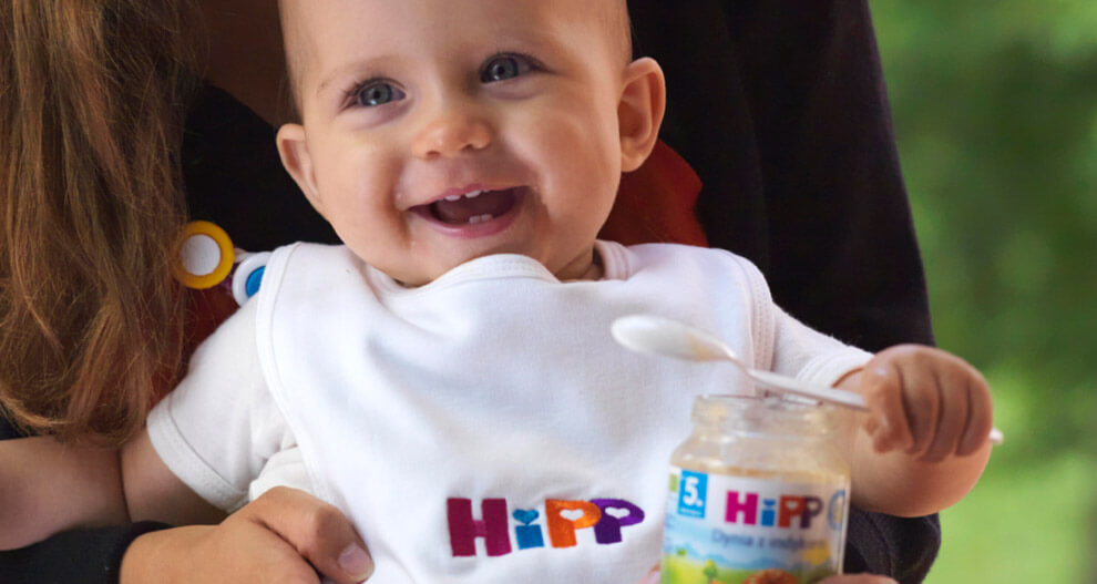 A baby wearing a white bib with the word "HiPP" embroidered on it is being fed from a jar of HiPP baby food.