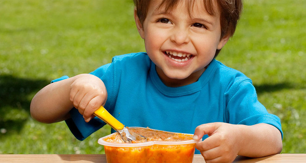 A child in a blue shirt is eating from a container with a yellow spoon.