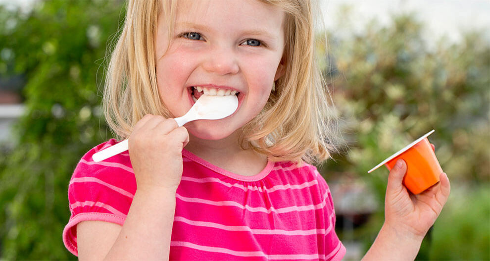 A young girl with blonde hair is eating from a small orange container with a white spoon.