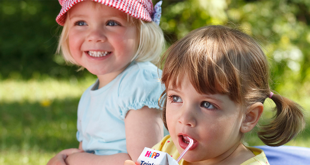 Two young girls sit outdoors, one wearing a red and white hat, the other sipping from a white box.