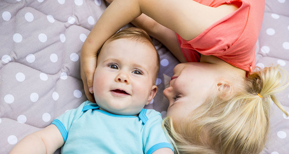 Two children, a girl and a baby, are lying on a gray bed with white polka dots.