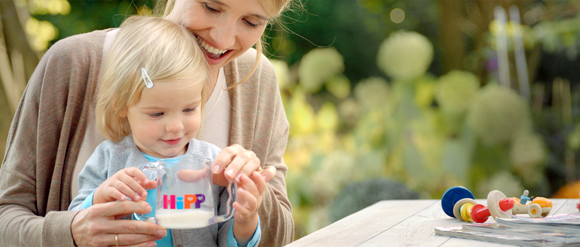 Una donna e un bambino sono all'aperto e il bambino tiene in mano una tazza trasparente con la scritta "HiPP".