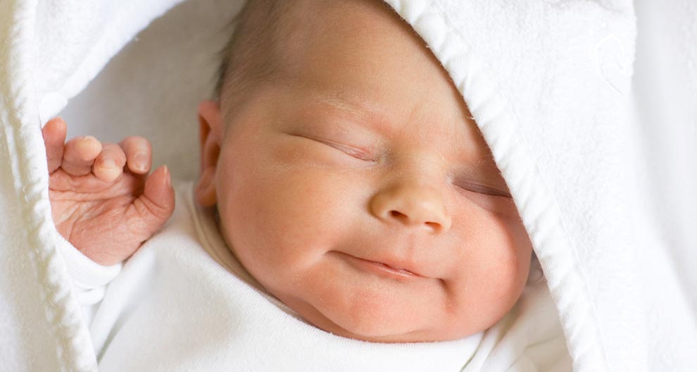 Primo piano del volto di un neonato, parzialmente coperto da un panno bianco, con una mano alzata.